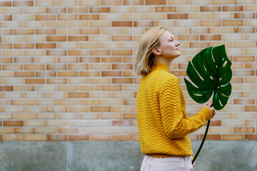 Smiling woman holding monstera leaf in front of brick wall. Concept of Ecology, Sustainability.