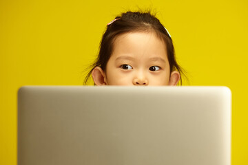 Pensive Asian Preschool Girl with a big opened eyes Engaged in Distance Learning via Laptop sitting against yellow isolated background