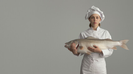 An apprentice chef in white attire grips an enormous fish carefully, highlighting her skill