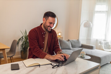 A smiling young adult businessman working from home typing on a laptop