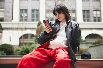 Young woman in stylish gray jacket and red pants sitting on bench with mobile phone against urban...