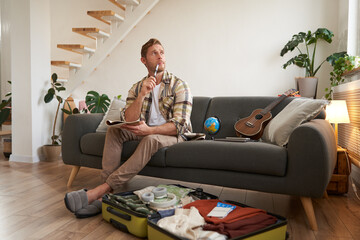Portrait of man thinking what to take with him on vacation, holding notebook, writing list of items with thoughtful face, preparing to go on holiday, sitting near suitcase with clothes - obrazy, fototapety, plakaty