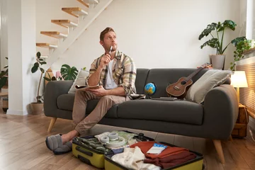 Gartenposter Portrait of man thinking what to take with him on vacation, holding notebook, writing list of items with thoughtful face, preparing to go on holiday, sitting near suitcase with clothes © Mix and Match Studio