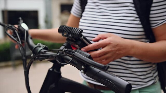 Close-up woman's hands checking mobile app on smartphone, setting up a route on internet online map navigator while riding electric bike in city. Close-up female cyclist using mobile phone in the city