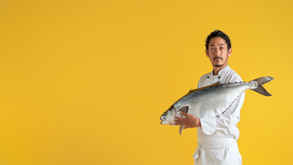 Confident male chef in white uniform holding a sizable fresh fish, ready for culinary preparation, against a yellow background - obrazy, fototapety, plakaty