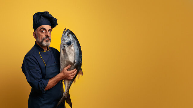 Experienced chef in dark attire giving a sly glance while presenting a large fish, set against a plain yellow background