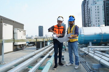 Foreman engineer and contractor wearing reflective jacket, engineering helmet, holding blueprints and walkie-talkie, inspects construction work and plumbing utilities on building rooftops.