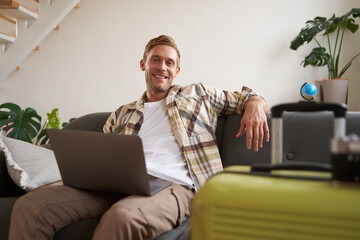 Portrait of happy young man, tourist booking his holiday with travel agency, looking at laptop, packed suitcase to go on vacation - obrazy, fototapety, plakaty