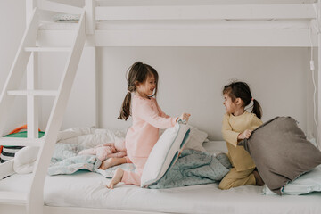 Adorable little girls have a pillow fight at home on the bed. Happy sisters are fighting with pillows. - 767169786
