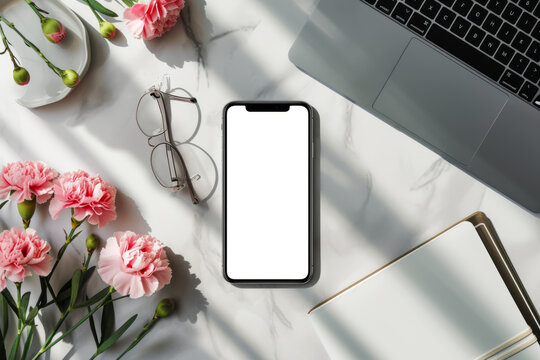 Mobile phone with white screen mockup with space for text or inscriptions on beautiful desk background with laptop, flowers, glasses, and blank notebook, top view
