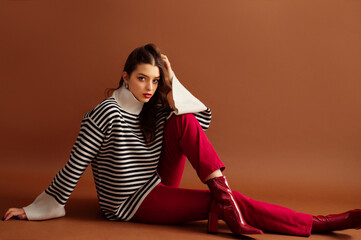Fashionable confident woman wearing stylish striped woolen sweater, red jeans, patent leather  ankle boots, posing on brown background. Studio fashion portrait. Copy, empty, blank space for text
