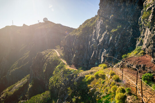 The picture depicts the picturesque landscape of Madeira. A path winds through rocky hills covered in lush greenery. The sun gently illuminates the scene, creating a golden glow and accentuating the 