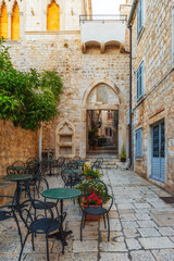 Fototapeta na wymiar Cafe tables and chairs outside in old cozy street in the in old medieval town Hvar in outdoor restaurant with nobody, Dalmatia, Croatia. Vertical orientation. Popular travel and tourist destination