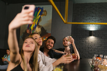 Young blonde women take selfie with cocktails in hand background male visitors of bar. Young people at bar counter chatter and get to know each other better - obrazy, fototapety, plakaty