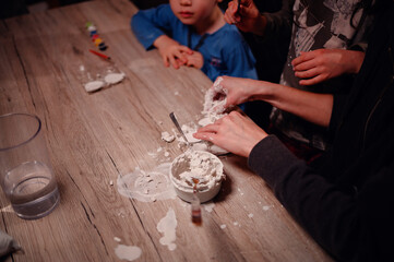A young boy and his sister focus intently on a science experiment at their home table, guided by a helpful woman, showcasing early learning and family collaboration