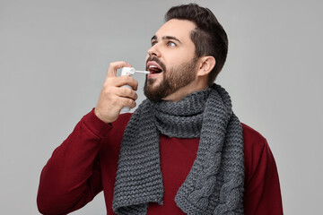 Young man with scarf using throat spray on grey background
