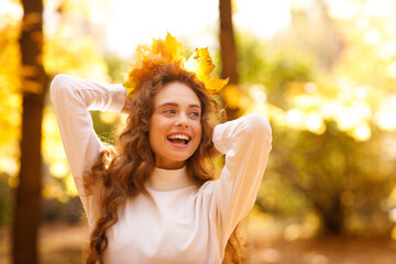 Smiling young woman with the fall crown of yellow leaves on her curly hairstyle head in the autumn park at sunset - 767211576
