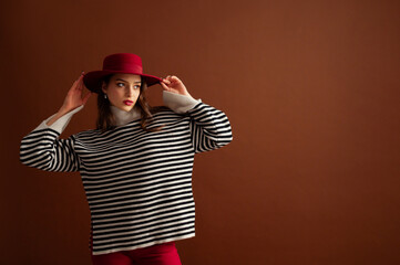 Fashionable confident woman wearing oversized striped knitted turtleneck sweater with wide sleeves, red hat, posing on brown background. Studio fashion portrait. Copy, empty, blank space for text - 767211967