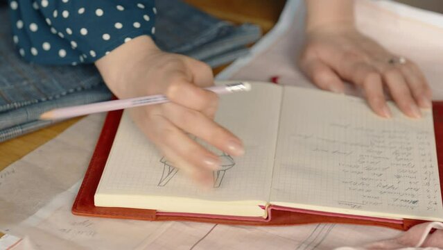 Close-up of a female fashion designer's hands as she sketches a new dress design on her notepad amidst various patterns and fabrics.