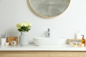 Vase with beautiful white roses and toiletries near sink in bathroom