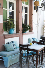 Street Cafe with Plants and Cozy Bench Seating. A street cafe corner with a cozy bench, striped cushions, wooden tables and chairs, and potted plants by the windows.