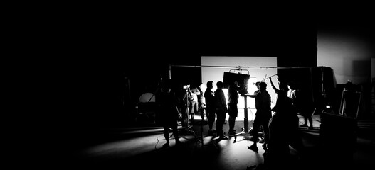 Silhouette images of video production behind the scenes of making of TV commercial movie shoot that film crew team lightman and camera man working together with director and equipment in big studio.
