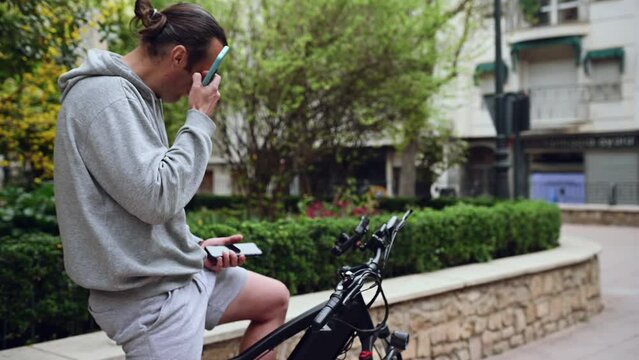 Young man setting up adjustments on smartphone, sitting on his electric bike, before riding in the city. Guy using mobile phone, renting an electric bike using a rental app. Bike sharing city service