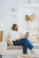 Smiling woman in glasses sitting on sofa at home, relaxed using laptop, working remotely online,...