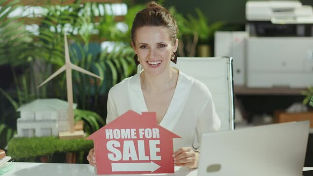 Sustainable real estate business. smiling modern female real estate agent in modern green office in white blouse with home for sale sign and laptop.
