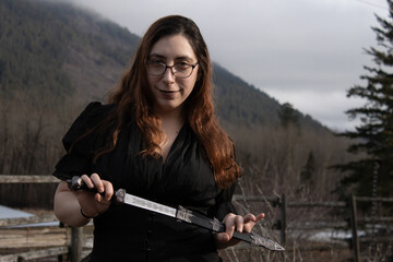 Beautiful High Fantasy Goth Witchy Cosplay Game Master LARP Halloween Long Dark Haired Brunette Woman Wearing Glasses and Holding Dagger with Misty Mountain Background with Space for Text