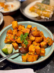 Close-up of seasoned sweet potatoes in cube form, garnished with herbs on a ceramic plate, accompanied by a slice of lime.