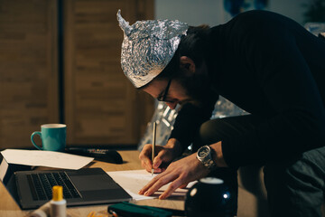 Conspiracy theory. A man in a tinfoil hat looks for signs while watching a video on a laptop.