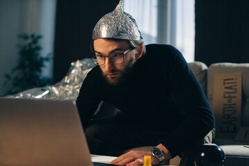 Conspiracy theory. A man in a tinfoil hat looks for signs while watching a video on a laptop. - obrazy, fototapety, plakaty