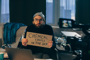 A conspiracy theorist shoots pseudoscientific videos on camera. A man in a tinfoil hat and a sign in his hands sits on a couch in front of the camera. - obrazy, fototapety, plakaty