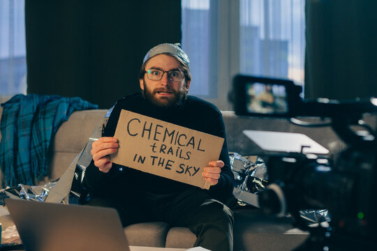 A conspiracy theorist shoots pseudoscientific videos on camera. A man in a tinfoil hat and a sign in his hands sits on a couch in front of the camera.