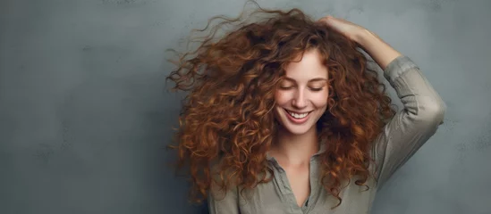 Fotobehang A happy woman with red curly Jheri curl hair is smiling while touching her ringlet hair. She looks joyful and her artificial hair integration adds volume to her surfer hair © AkuAku