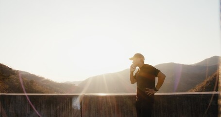 A man is standing on a ledge looking out over a mountain range. He is wearing a black shirt and a hat