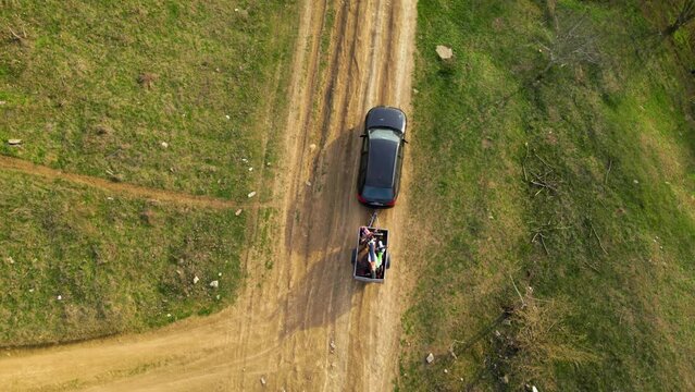 Aerial high angle top down shot of a black car towing a small trailer with two off road cross morotbikes along a windy dirt road. Drone footage of vehicle with a trailer behind going to sports event.
