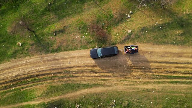 Aerial high angle top down shot of a black car towing a small trailer with two off road cross morotbikes along a windy dirt road. Drone footage of vehicle with a trailer behind going to sports event.
