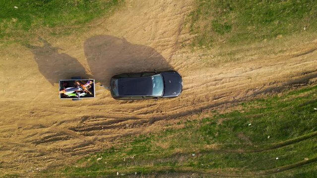 Aerial high angle top down shot of a black car towing a small trailer with two off road cross morotbikes along a windy dirt road. Drone footage of vehicle with a trailer behind going to sports event.
