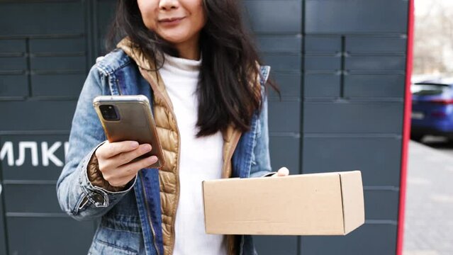 Woman receiving parcel from post terminal machine using smartphone outdoors. Parcel delivery machine. Mail delivery and post service, online shopping, e commerce concept.