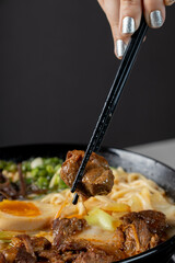 A hand picking up a piece of ramen meat with chopsticks close up