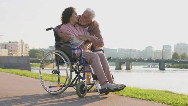 Senior woman in wheelchair walking with caregiver old man on road in park. Elderly family couple man supporting embracing paralyzed woman in chair for people with disability outdoor. Rehabilitation