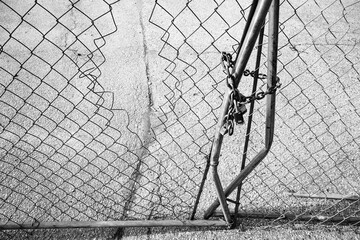 A black and white image of a damaged, metal chain link fence and locked gate
