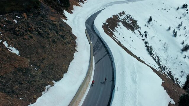 Aerial footage, camera fly over two cyclists ride on empty secluded road on a mountain. Cycling in tough cold winter conditions, snow walls on road sides. Cycling in winter