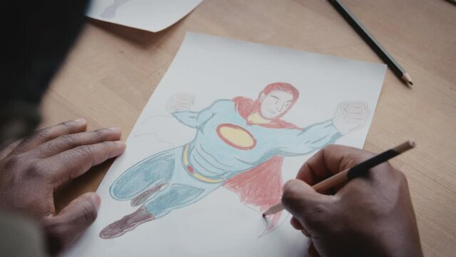 Close up over the shoulder shot of male artist drawing superhero on paper with colored pencil while working at desk
