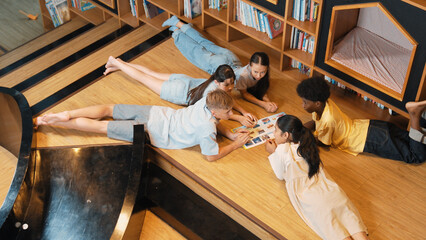 Group of diverse children lying down in circle while reading a book at library. Top view of girl...