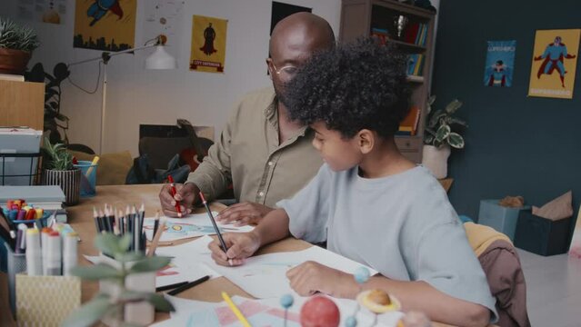African American father and little son sitting at desk in kids room at home and drawing together with colored pencils on paper