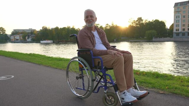 Happy senior man in wheelchair smiling enjoying freedom walking on road in city park outdoor. Old cheerful positive man in chair for people with disability. Disability disease rehabilitation