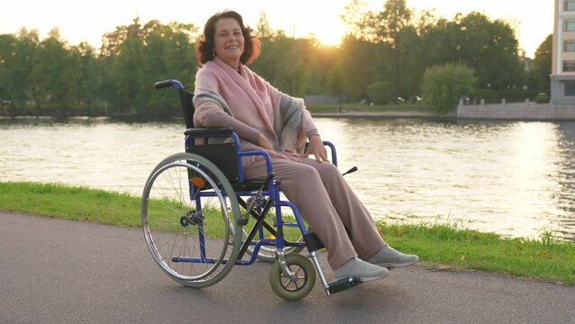 Happy senior woman in wheelchair smiling enjoying freedom walking on road in city park outdoor. Old cheerful positive woman in chair for people with disability. Disability disease rehabilitation